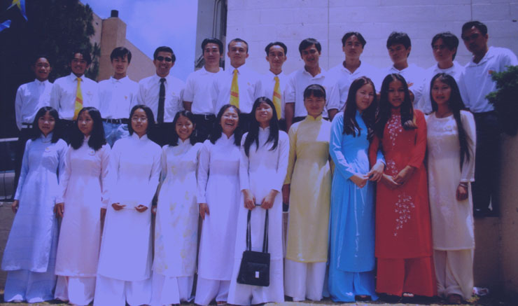 Foundation members posing together outside a building: young women in white, blue, red and gold áo dài in front, young men in white shirts and yellow ties behind